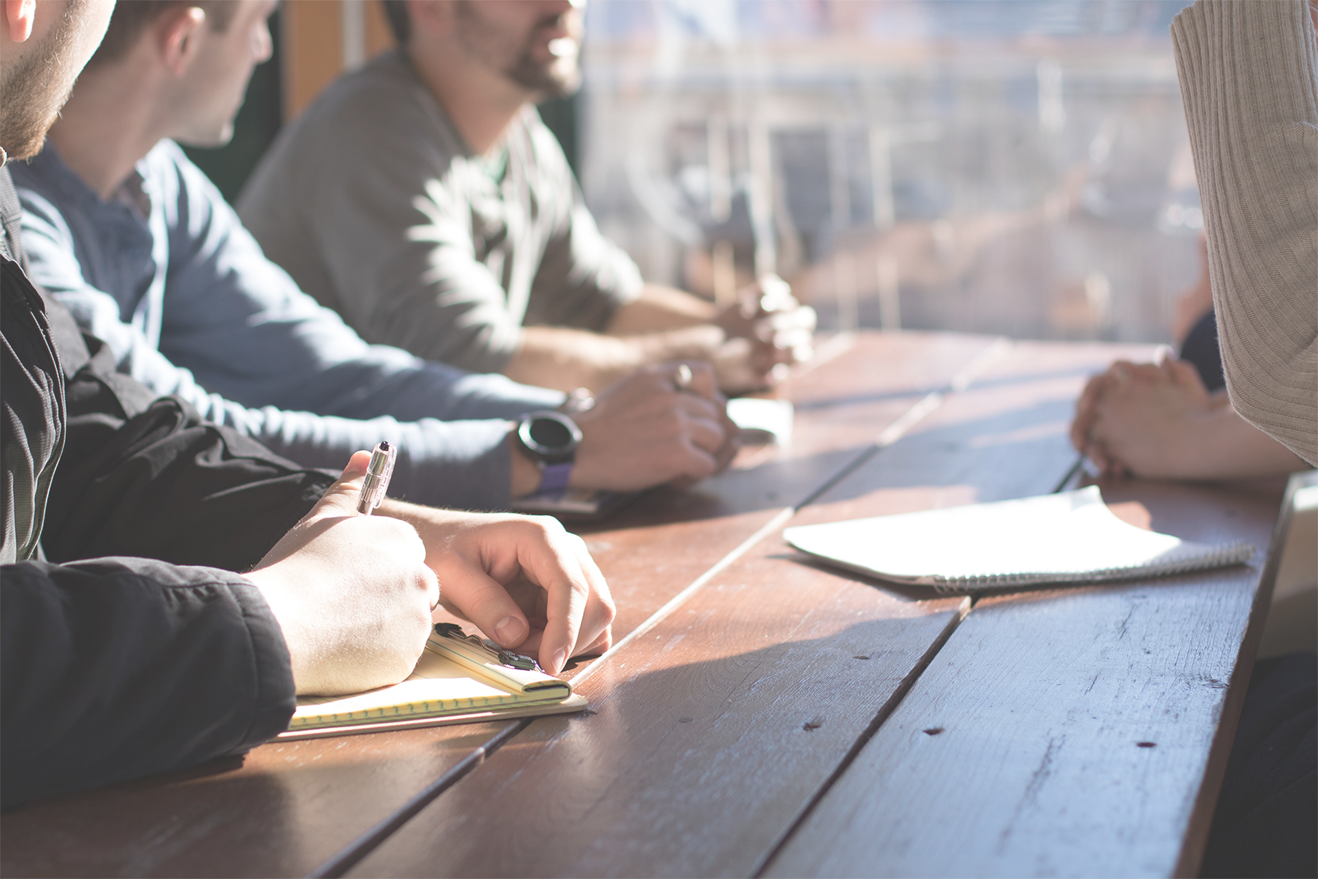 Group of people meeting on a wooden table with a window in the background. The view from the window is blurred.